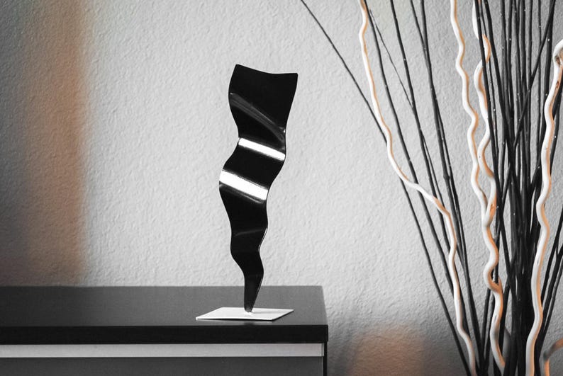 May include: A black metal sculpture with a wavy, abstract design. The sculpture is standing on a small, square base.