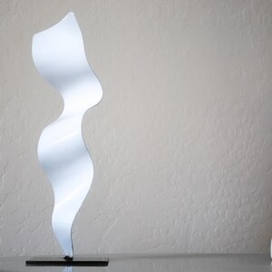 May include: White abstract metal sculpture with a wavy, flowing design. The sculpture is on a black base.