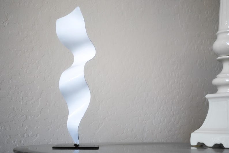 May include: White abstract metal sculpture with a wavy, flowing design. The sculpture is on a black base.