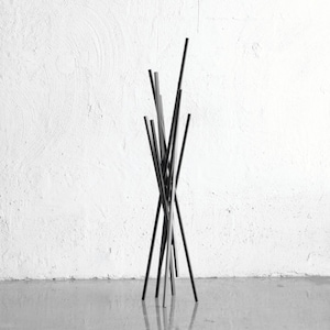 May include: A black metal coat rack with a unique, abstract design. The rack is made of thin, vertical rods that are arranged in a random pattern. The rack is standing on a concrete floor in front of a white wall.
