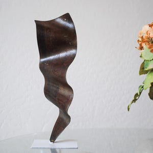 May include: A dark brown metal sculpture with a wavy, abstract design. The sculpture is standing on a white base.