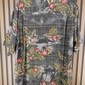 May include: Gray short-sleeve shirt with a tropical floral print. The shirt features a pattern of red hibiscus flowers and green leaves on a gray background with a subtle plaid design. The shirt has a classic collar and short sleeves.