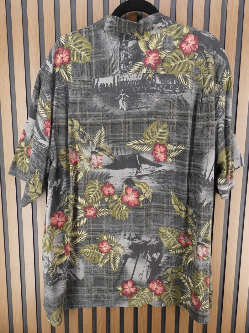 May include: Gray short-sleeve shirt with a tropical floral print. The shirt features a pattern of red hibiscus flowers and green leaves on a gray background with a subtle plaid design. The shirt has a classic collar and short sleeves.