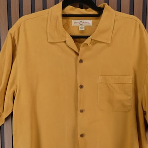 May include: Mustard yellow short-sleeve button-down shirt with a chest pocket. The shirt has a collared neckline and a label with text. The shirt is hanging on a black hanger.
