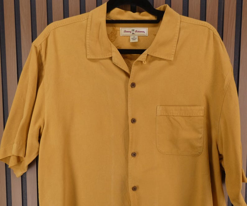 May include: Mustard yellow short-sleeve button-down shirt with a chest pocket. The shirt has a collared neckline and a label with text. The shirt is hanging on a black hanger.