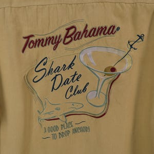 May include: Yellow button-up shirt with embroidered design. The design includes the text "Tommy Bahama Shark Date Club", a martini glass with an olive, and a shark. The phrase "A good place to drop anchor!" is also included.