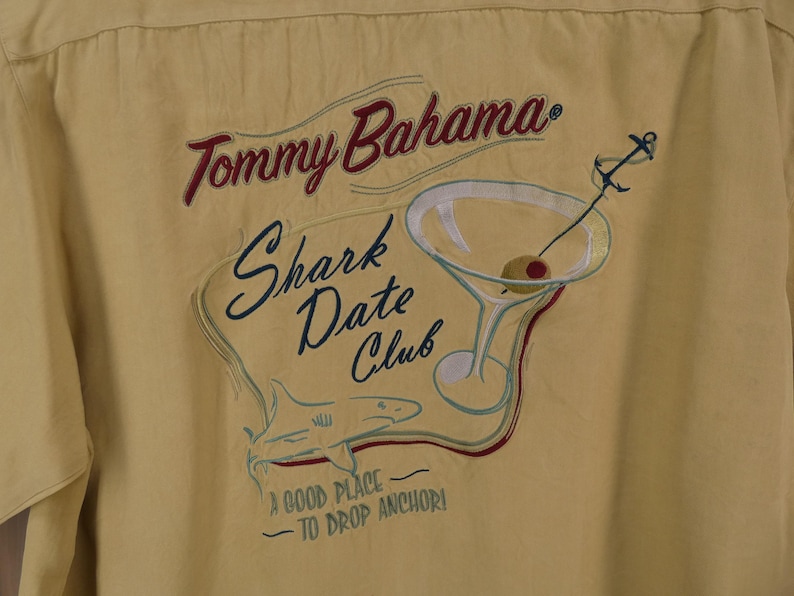 May include: Yellow button-up shirt with embroidered design. The design includes the text "Tommy Bahama Shark Date Club", a martini glass with an olive, and a shark. The phrase "A good place to drop anchor!" is also included.