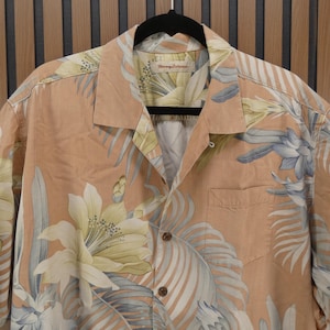 Puede incluir: Camisa abotonada color melocot&oacute;n con un dise&ntilde;o floral en blanco y azul. La camisa, colgada de una percha negra, tiene un bolsillo en el pecho izquierdo. El dise&ntilde;o presenta grandes flores y hojas tropicales.