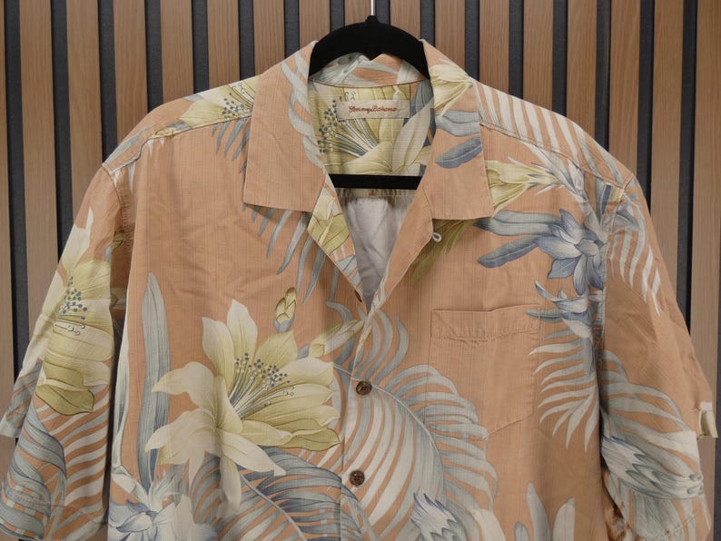 Puede incluir: Camisa abotonada color melocot&oacute;n con un dise&ntilde;o floral en blanco y azul. La camisa, colgada de una percha negra, tiene un bolsillo en el pecho izquierdo. El dise&ntilde;o presenta grandes flores y hojas tropicales.