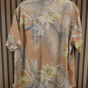 Puede incluir: Una camisa de manga corta con un estampado floral tropical. La camisa tiene un fondo color melocot&oacute;n con un dise&ntilde;o de flores blancas, amarillas y azules y hojas de palma. La camisa est&aacute; colgada de una percha negra.