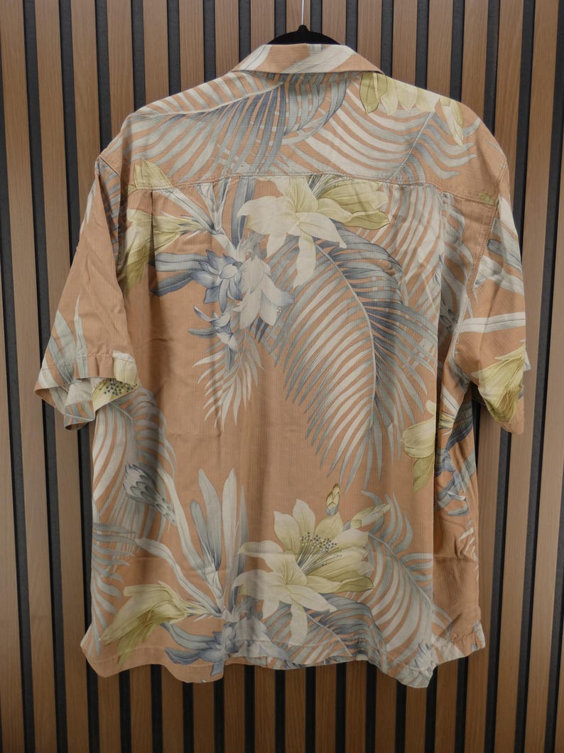 Puede incluir: Una camisa de manga corta con un estampado floral tropical. La camisa tiene un fondo color melocot&oacute;n con un dise&ntilde;o de flores blancas, amarillas y azules y hojas de palma. La camisa est&aacute; colgada de una percha negra.