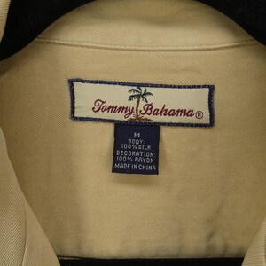 May include: Close-up of a tan Tommy Bahama shirt. The shirt's label is visible, featuring the brand name and a palm tree logo. The label also indicates the shirt is size M, made of 100% silk, with rayon decoration, and made in China.