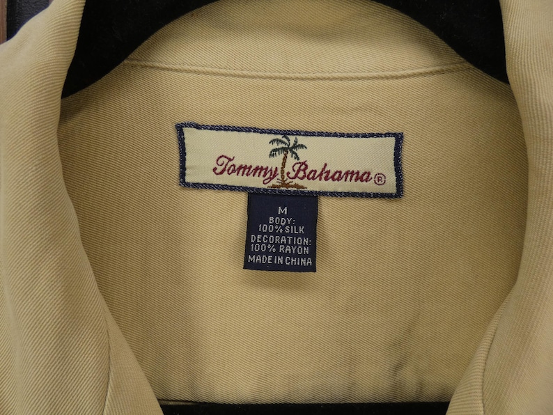May include: Close-up of a tan Tommy Bahama shirt. The shirt's label is visible, featuring the brand name and a palm tree logo. The label also indicates the shirt is size M, made of 100% silk, with rayon decoration, and made in China.