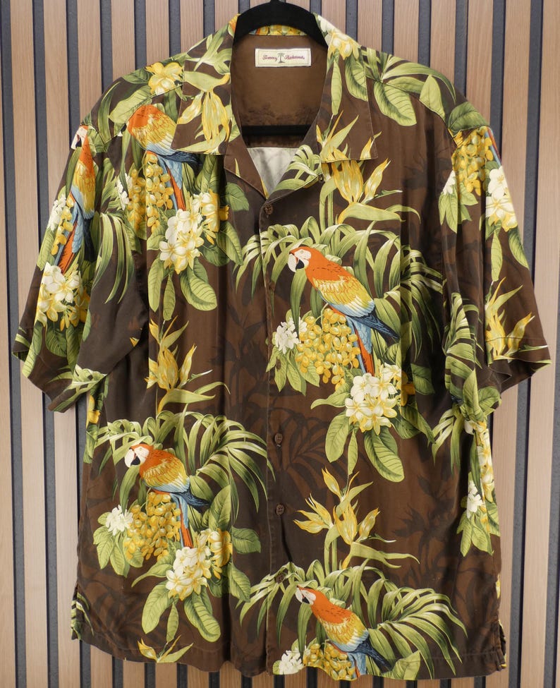 May include: Brown button-down shirt with a tropical parrot and floral print. The short-sleeved shirt features a vibrant design with parrots, yellow flowers, and green leaves. The shirt has a classic collar and is made of a lightweight fabric.