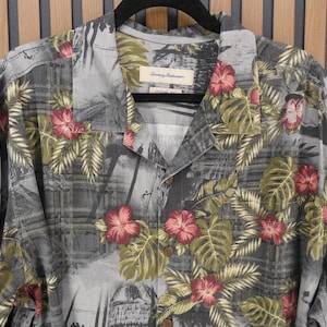 May include: A short-sleeved button-down shirt with a tropical floral print. The shirt features a gray background with a pattern of red hibiscus flowers and green leaves. The shirt is hanging on a black hanger.