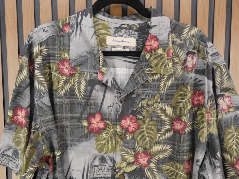May include: A short-sleeved button-down shirt with a tropical floral print. The shirt features a gray background with a pattern of red hibiscus flowers and green leaves. The shirt is hanging on a black hanger.