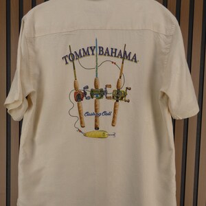 May include: Cream-colored short-sleeved shirt with a graphic of fishing rods and the text "Tommy Bahama" and "Casting Call". The graphic features three fishing rods with reels and a yellow lure.