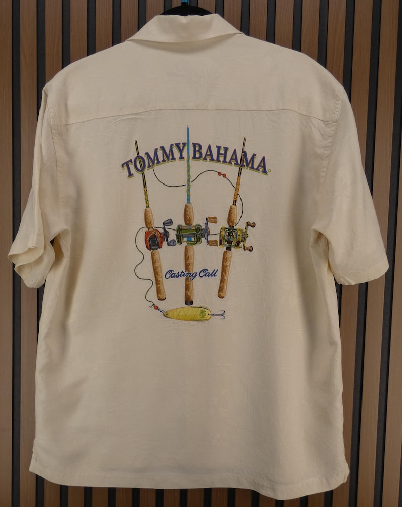 May include: Cream-colored short-sleeved shirt with a graphic of fishing rods and the text "Tommy Bahama" and "Casting Call". The graphic features three fishing rods with reels and a yellow lure.