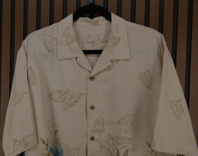 K&ouml;nnte beinhalten: Cremefarbenes Button-Down-Hemd mit Bl&auml;ttermuster. Das Hemd hat kurze &Auml;rmel und einen klassischen Kragen. Der Markenname "Tommy Bahama" ist auf dem Kragen gestickt. Das Hemd ist aus leichtem Stoff, ideal f&uuml;r warmes Wetter.