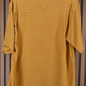 May include: Mustard yellow short-sleeved shirt with a collared neckline. The shirt has a button-down front and a relaxed fit. The back of the shirt features a yoke detail. The shirt is hanging on a black hanger.