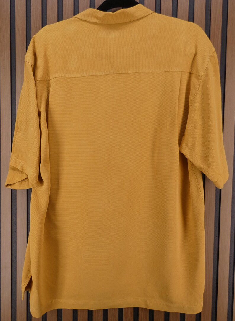 May include: Mustard yellow short-sleeved shirt with a collared neckline. The shirt has a button-down front and a relaxed fit. The back of the shirt features a yoke detail. The shirt is hanging on a black hanger.