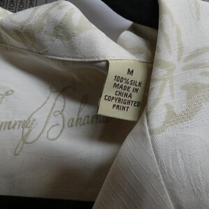 K&ouml;nnte beinhalten: Nahaufnahme eines cremefarbenen Seidenhemdes mit dezentem Blumenmuster. Das Hemd hat das gestickte "Tommy Bahama"-Logo und ein Etikett mit der Aufschrift "100% Silk Made in China Copyrighted Print".