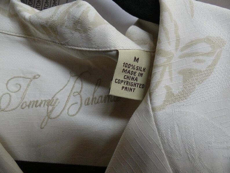 K&ouml;nnte beinhalten: Nahaufnahme eines cremefarbenen Seidenhemdes mit dezentem Blumenmuster. Das Hemd hat das gestickte "Tommy Bahama"-Logo und ein Etikett mit der Aufschrift "100% Silk Made in China Copyrighted Print".