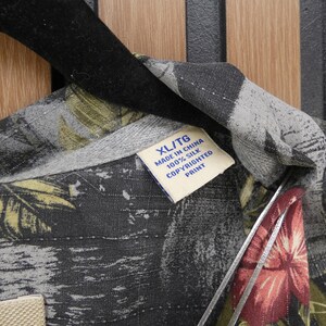 May include: Close-up of a silk shirt with a tropical floral print in shades of gray, green, and red. The shirt has a collar and a tag that reads "XL/TG MADE IN CHINA 100% SILK COPYRIGHTED PRINT."