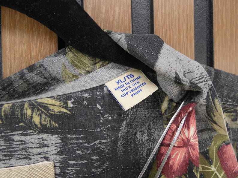 May include: Close-up of a silk shirt with a tropical floral print in shades of gray, green, and red. The shirt has a collar and a tag that reads "XL/TG MADE IN CHINA 100% SILK COPYRIGHTED PRINT."