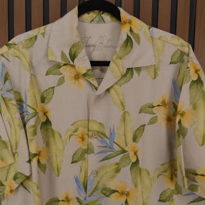 May include: A button-down shirt with a floral pattern. The shirt is beige with yellow flowers, green leaves, and blue accents. The brand name "Tommy Bahama" is visible on the collar.