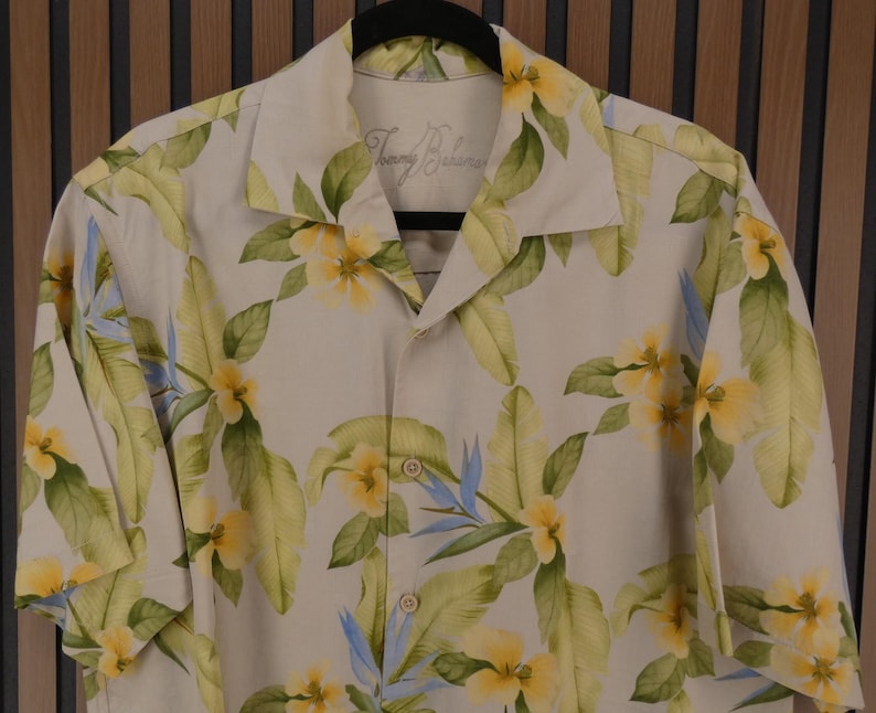 May include: A button-down shirt with a floral pattern. The shirt is beige with yellow flowers, green leaves, and blue accents. The brand name "Tommy Bahama" is visible on the collar.