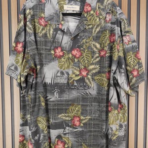 May include: Gray short-sleeve button-down shirt with a tropical floral print. The shirt features a pattern of red hibiscus flowers and green leaves on a gray background. The shirt is hanging on a black hanger.