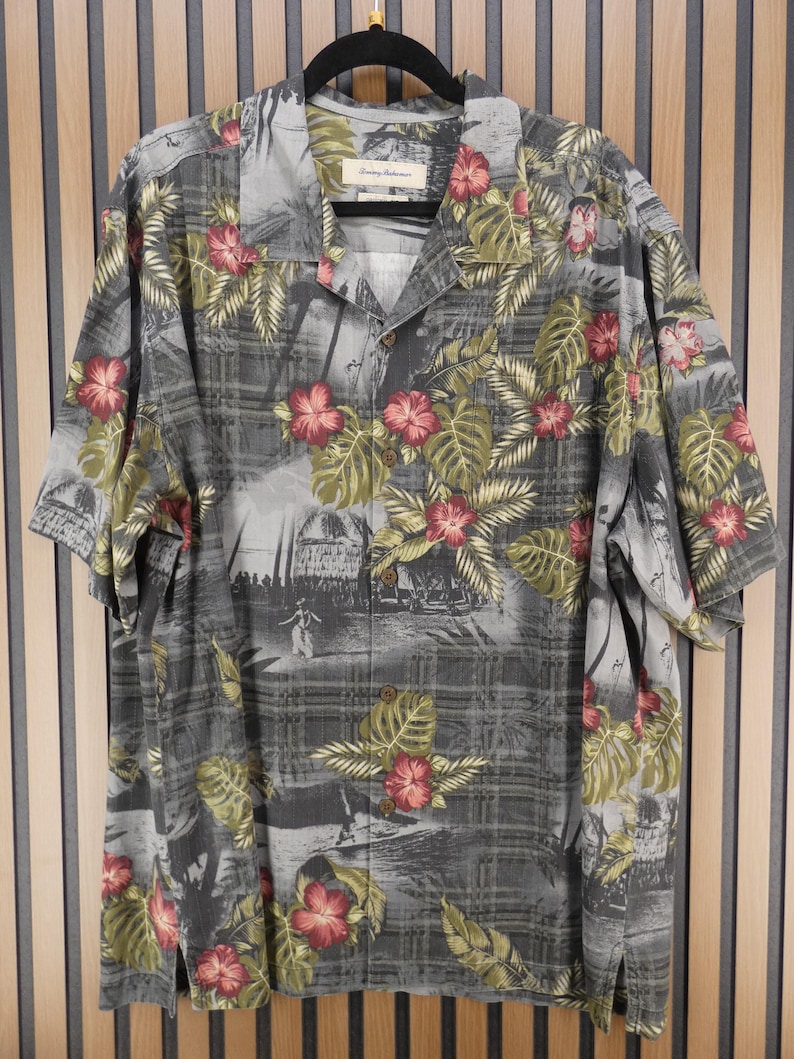 May include: Gray short-sleeve button-down shirt with a tropical floral print. The shirt features a pattern of red hibiscus flowers and green leaves on a gray background. The shirt is hanging on a black hanger.
