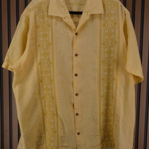 May include: A pale yellow short-sleeved shirt with embroidered floral designs down the front. The shirt features a collared neckline and wooden buttons. The brand name "Tommy Bahama" is visible on the collar.