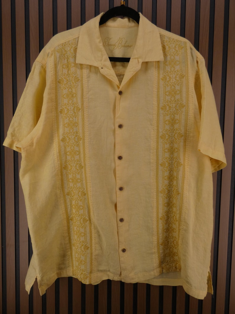 May include: A pale yellow short-sleeved shirt with embroidered floral designs down the front. The shirt features a collared neckline and wooden buttons. The brand name "Tommy Bahama" is visible on the collar.