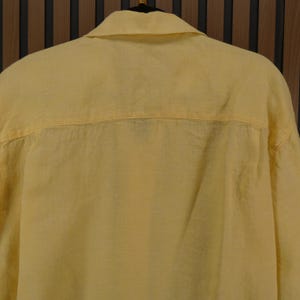 May include: A pale yellow button-down shirt made from a lightweight material. The shirt features a classic collar and a relaxed fit, suitable for casual wear. The back of the shirt is visible, showing the yoke and the fabric's texture.