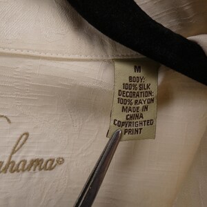 May include: Close-up of a cream-colored shirt tag. The tag reads "M" and lists the fabric composition as 100% silk for the body and 100% rayon for the decoration. The shirt is made in China and has a copyrighted print.