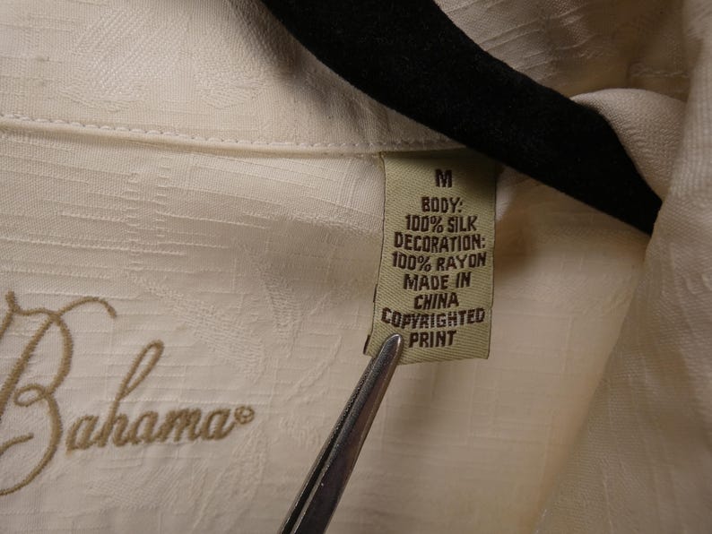 May include: Close-up of a cream-colored shirt tag. The tag reads "M" and lists the fabric composition as 100% silk for the body and 100% rayon for the decoration. The shirt is made in China and has a copyrighted print.