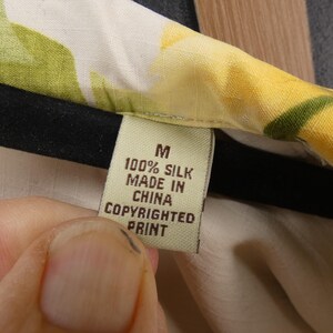 May include: Close-up of a garment tag. The tag is beige with black text that reads "M 100% SILK MADE IN CHINA COPYRIGHTED PRINT". The garment has a floral pattern with yellow and green hues.
