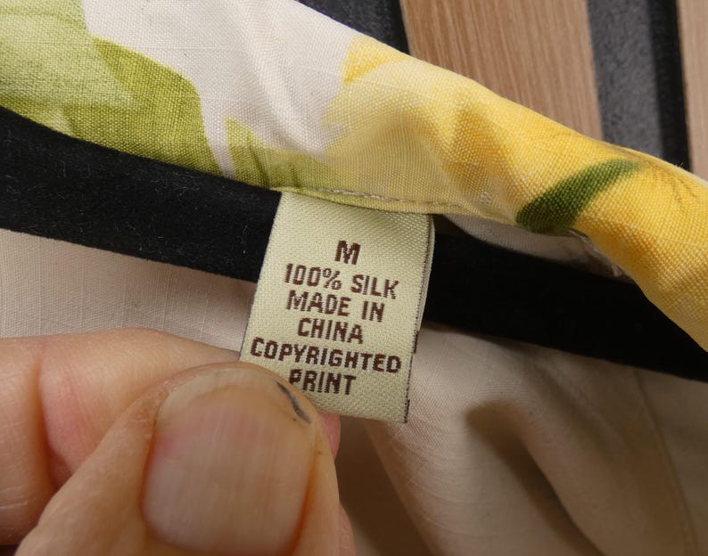 May include: Close-up of a garment tag. The tag is beige with black text that reads "M 100% SILK MADE IN CHINA COPYRIGHTED PRINT". The garment has a floral pattern with yellow and green hues.