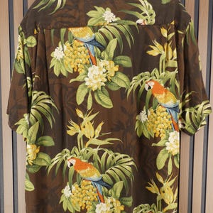May include: Brown short-sleeve shirt with a tropical print featuring parrots, yellow flowers, and green leaves. The shirt has a button-down collar and is made of a lightweight fabric. The design includes a repeating pattern of parrots and floral elements.