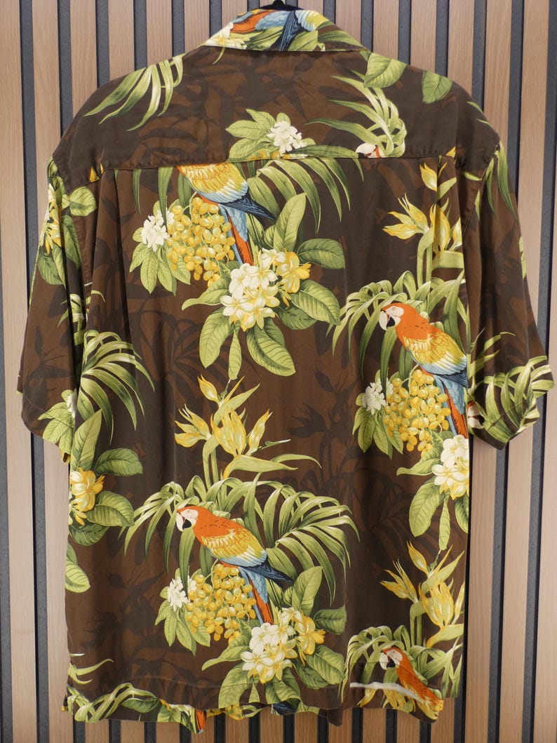 May include: Brown short-sleeve shirt with a tropical print featuring parrots, yellow flowers, and green leaves. The shirt has a button-down collar and is made of a lightweight fabric. The design includes a repeating pattern of parrots and floral elements.