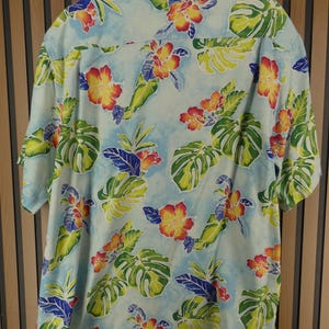 May include: Light blue short-sleeve shirt with a tropical floral pattern. The shirt features large green leaves, red and yellow flowers, and blue accents. The shirt is made of a lightweight fabric.