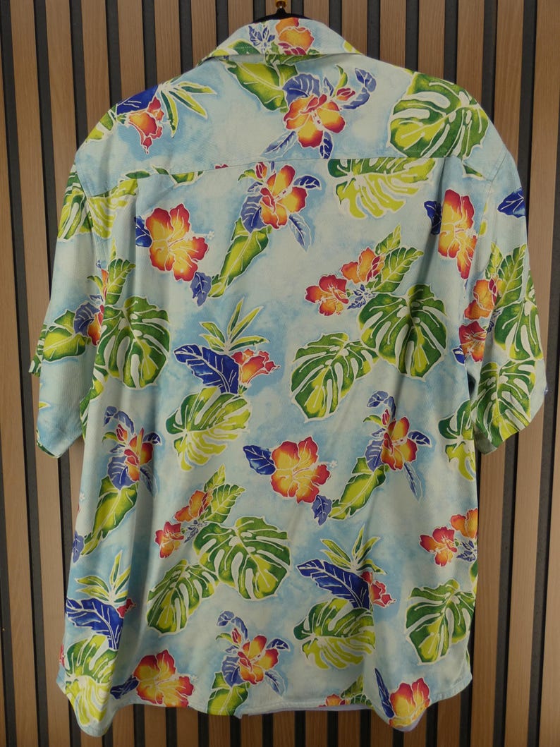 May include: Light blue short-sleeve shirt with a tropical floral pattern. The shirt features large green leaves, red and yellow flowers, and blue accents. The shirt is made of a lightweight fabric.