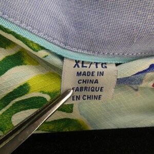 May include: Close-up of a tag on a patterned shirt. The tag reads "XL/TG MADE IN CHINA FABRIQUE EN CHINE". The shirt has a light blue collar and a colorful floral pattern with green and yellow leaves.