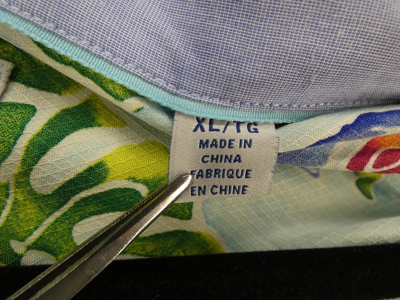 May include: Close-up of a tag on a patterned shirt. The tag reads "XL/TG MADE IN CHINA FABRIQUE EN CHINE". The shirt has a light blue collar and a colorful floral pattern with green and yellow leaves.