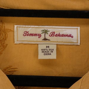 May include: Mustard yellow silk shirt with a textured pattern and embroidered details. The label reads "Tommy Bahama" with a palm tree logo. The shirt is labeled as size M and made in China.