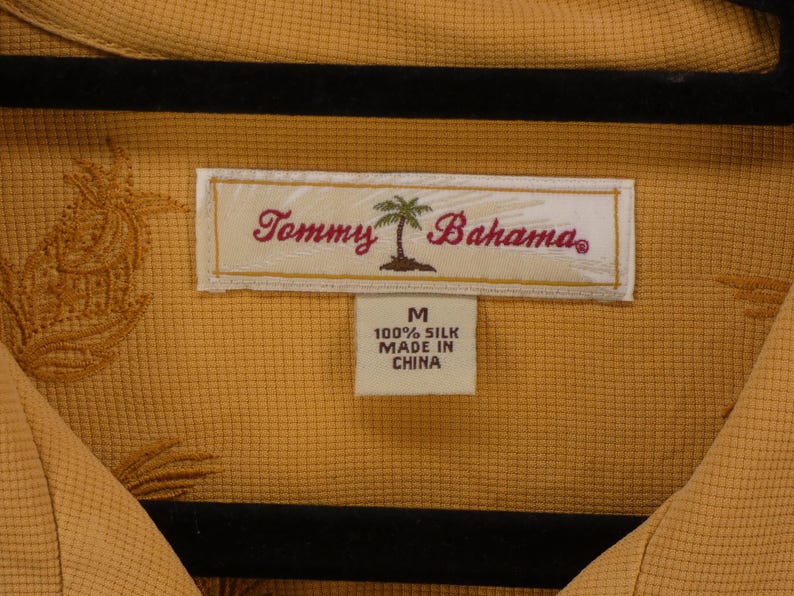 May include: Mustard yellow silk shirt with a textured pattern and embroidered details. The label reads "Tommy Bahama" with a palm tree logo. The shirt is labeled as size M and made in China.