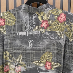 May include: Gray button-up shirt with a tropical floral and landscape print. The shirt features red hibiscus flowers, green leaves, and a grayscale scene. The shirt is hanging on a black hanger.