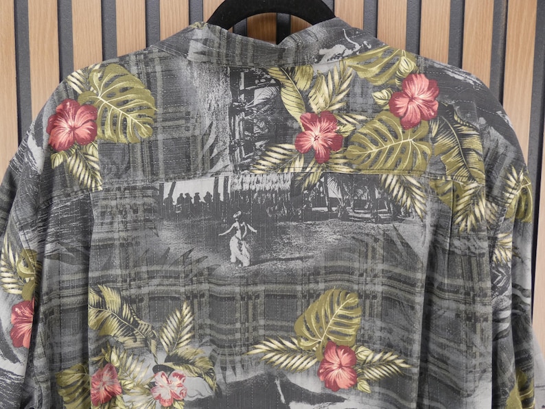 May include: Gray button-up shirt with a tropical floral and landscape print. The shirt features red hibiscus flowers, green leaves, and a grayscale scene. The shirt is hanging on a black hanger.